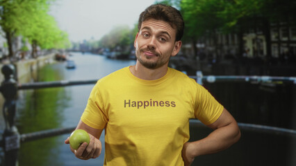 Man holding green apple on a street bridge in amsterdam wearing yellow shirt with 'happiness' print; happiness healthy lifestyle.