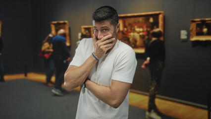 Man with hand covering mouth and crossed arms in a building gallery observing framed paintings;...