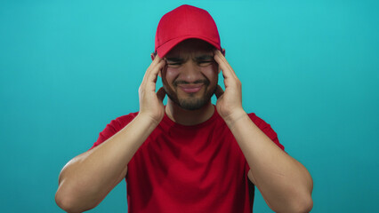 Man wearing red cap and shirt pressing temples with both hands against turquoise studio wall; distress.