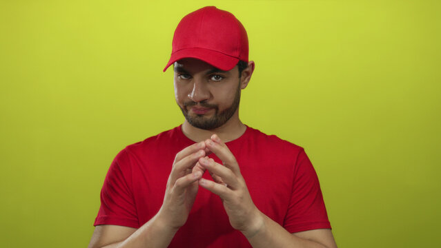 Hispanic courier man wearing red cap and shirt presses hands together in steeple gesture in yellow studio; cunning strategy plan.