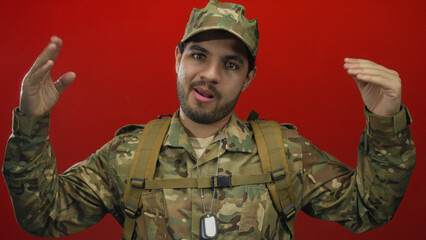 Man wearing camouflage uniform, backpack straps and dogtags gestures with both raised hands in...