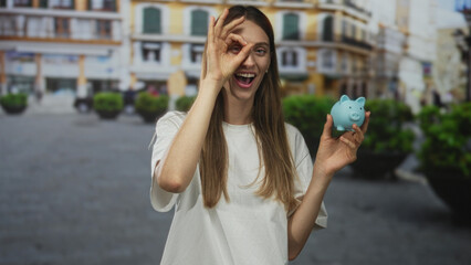 Woman holding blue piggy bank making ok sign over eye on a cobblestone street with large planters...