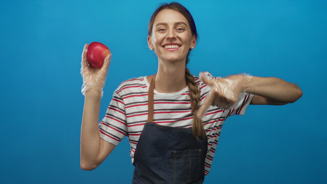 Woman wearing apron and disposable gloves holds red apple and points finger toward fruit in blue studio, striped shirt visible; cheerful.