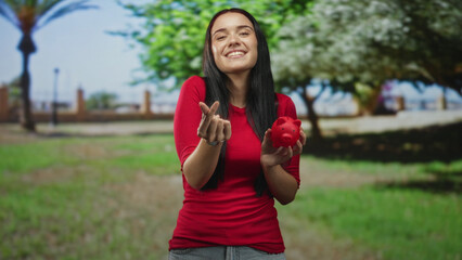 Woman holding red piggy bank and making finger heart gesture in park, smiling with closed eyes and...