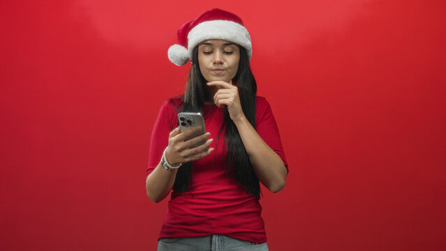 Fototapeta Young woman in santa hat holds smartphone, reads screen and touches chin against red backdrop in studio  thoughtful planning.