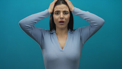 Woman expressing frustration with hands on head against a blue background, wearing a light blue...