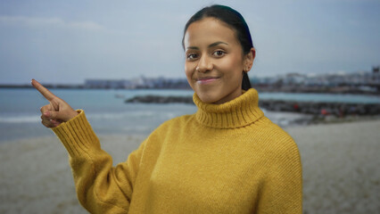 Woman in yellow sweater points at seaside on beach with cloudy sky, showcasing serene and calm coastal environment while smiling peacefully outdoors with ocean background.