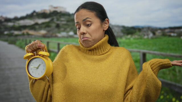 Woman in yellow sweater holds alarm clock in park expressing confusion with green field and cloudy sky in background.