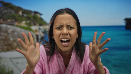 Woman in a pink bathrobe looking frustrated by the seaside with turquoise water and rocks, capturing an outdoors beach scene with a young hispanic woman.