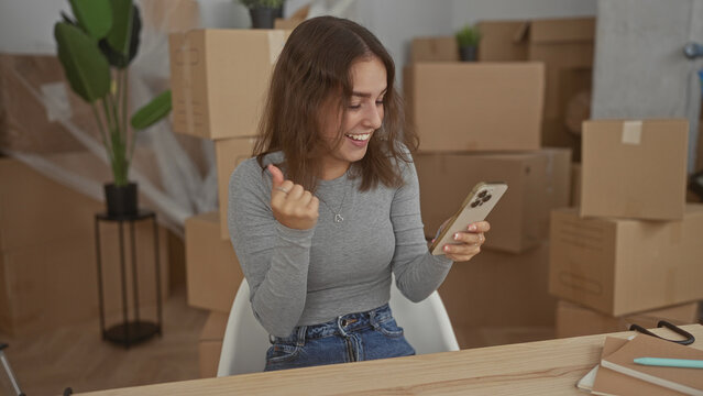 Woman sitting at wooden table holding smartphone with hands and fist pump in building among moving boxes; moving day joy. - Powered by Adobe