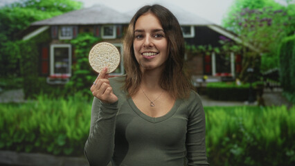Naklejka premium Woman holding ricecake between fingers at house entrance building, smiling while presenting the round snack to camera; healthy snack contentment.