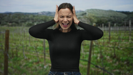 Fototapeta premium Woman in black top and jeans pressing hands to ears, eyes closed and mouth open in forest vineyard; emotional distress.