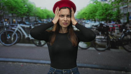 Woman wearing red beret pressing hands to temples with eyes closed for headache on street in amsterdam; anxiety overwhelm.