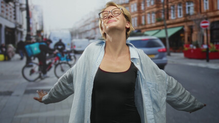 Woman with arms out exposing bare shoulder and neckline, smiling with glasses in open shirt on street with cyclists and cars nearby; carefree joy.