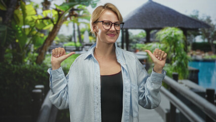 Fototapeta premium Woman pointing thumbs at herself on resort bridge building wearing glasses and casual shirt, smiling broadly with relaxed pose; confidence joy.