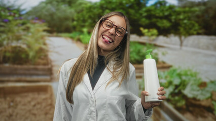 Woman holding a pump bottle in a greenhouse wearing a lab coat and glasses while smiling broadly; joy wellness.