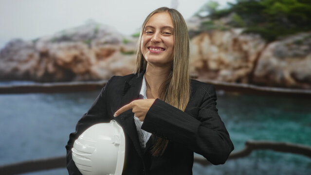 Young hispanic woman pointing finger to hardhat and holding helmet while smiling in a studio set with coastal backdrop; confidence safety. - Powered by Adobe