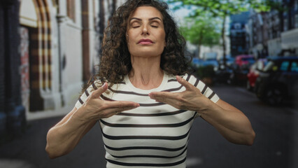 Young hispanic woman pressing both palms downward in calming gesture on bustling city street with parked cars visible; serenity.