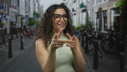Woman wearing green top and glasses smiling while pointing fingers at camera in city street; playful confidence.