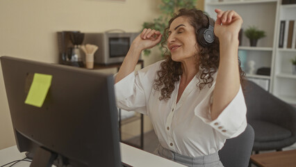 Woman raising arms dancing in office wearing headphones beside a computer monitor and keyboard on desk; pure joy.