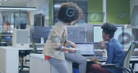 Perched woman sliding charts at laptop, seated peer typing and analyzing business data with HUD - Powered by Adobe