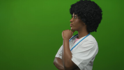 Naklejka premium African american woman doctor holds stethoscope while hand on chin in green studio; serious contemplation.
