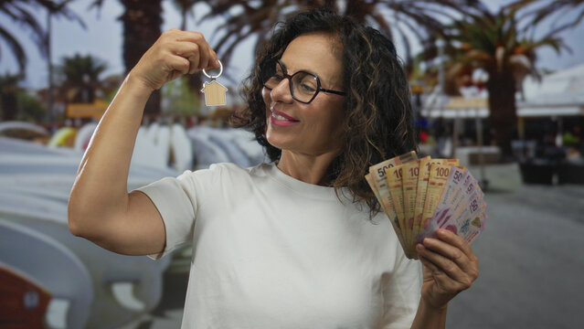 Middle-aged hispanic woman holds a house key and mexican pesos outdoors, symbolizing real estate and financial success in an urban setting.