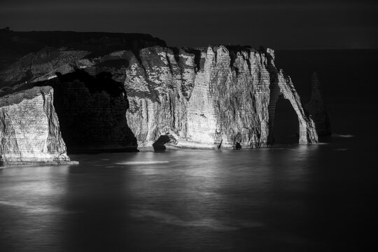 Dramatic illuminated sea cliffs at dusk in Étretat, Normandy, with glowing reflections on the calm coastal waters and the iconic natural rock arch formations.