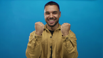 Fototapeta premium Man grins and clenches fists with broad smile in vibrant blue studio setting; victory celebration energy passion.