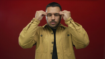 Young hispanic man wearing yellow jacket raises hands and opens mouth in red studio; frustration...