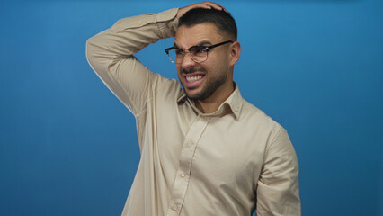 Young hispanic man adjusts glasses against a bright blue studio wall, squinting with a furrowed brow and tense posture; confusion.