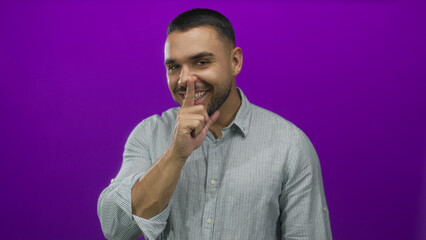 Young hispanic man smiling with finger to lips for silence in purple studio setting; mystery playfulness.