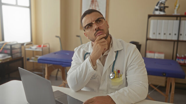 Young hispanic doctor with stethoscope has hand on chin while looking at laptop screen in clinic building; serenity.