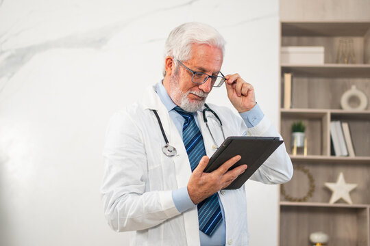 Mature senior male doctor in medical uniform using digital tablet in clinic. Therapist professional healthcare expert searching information online in hospital room. Medicine healthcare medical checkup