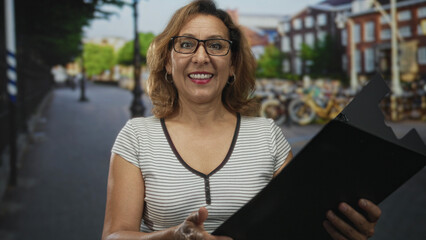 Woman holding black folder and reading intently on a street with parked bicycles and historic building facades; reflection.