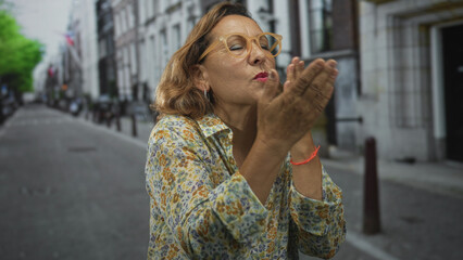 Woman blowing kiss with cupped hands on street, wearing glasses and floral shirt, palms visible and lips puckered; affection connection.