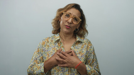 Woman clutches chest in studio set with light grey wall wearing patterned floral blouse, glasses and hoop earrings; gratitude.