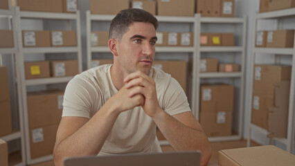 Man with parcel and arms crossed at desk while checking laptop among stacked boxes in storage...
