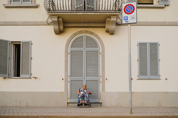 An elderly woman enjoys the outdoors, sitting in a wheelchair near a building with closed shutters. The setting captures a moment of tranquility and independence.