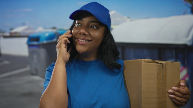Deliverywoman holds box with one hand and talks on phone on city street by large trash container; duty.