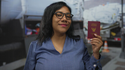 Woman wearing glasses smiles and holds thai passport while flexing bicep by baggage cart at airport; pride.