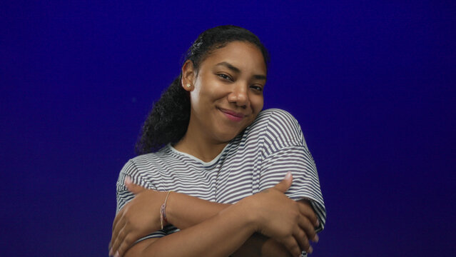 African american woman wearing striped shirt hugs arms with eyes closed in studio; self care and acceptance.