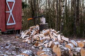 A chopping axe is lodged in a birch log on a tree stump, surrounded by a large pile of freshly split firewood next to a red wooden shed in a frosty forest clearing