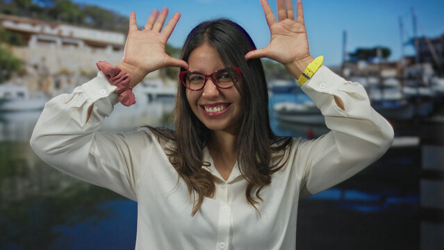Woman enjoys sunny seaside setting, smiling brightly near boats in a scenic port with a cheerful expression and stylish red glasses emphasizing youthful energy.