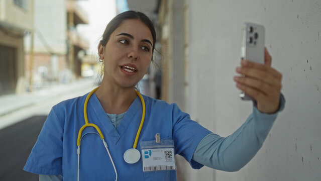 Young hispanic woman doctor wearing uniform and stethoscope holds smartphone for video call on street; telemedicine remote care empathy.