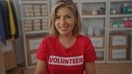 Blonde woman smiling while wearing a red volunteer shirt surrounded by packed boxes and supplies on shelves in a donation center; compassion generosity support.