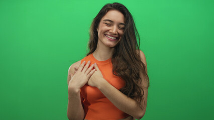 Young hispanic woman wearing orange sleeveless top with hands on chest gesture in green studio; gratitude sincerity.