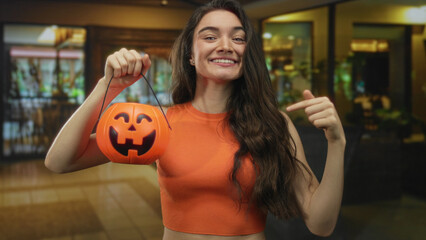 Woman holds halloween pumpkin bucket and points finger to object in brightly lit hotel lobby; excitement.