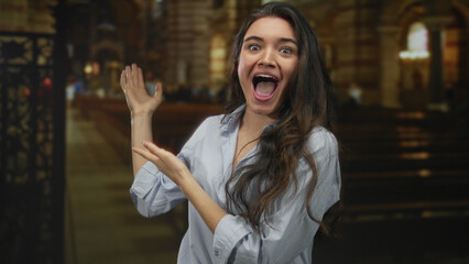 Young hispanic woman gestures with both hands while smiling inside a historic church building under...