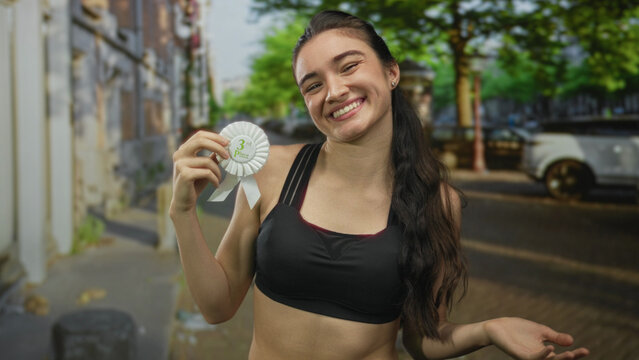 Woman smiling and holding a third place ribbon while shrugging shoulders on a busy city street; humble acceptance.